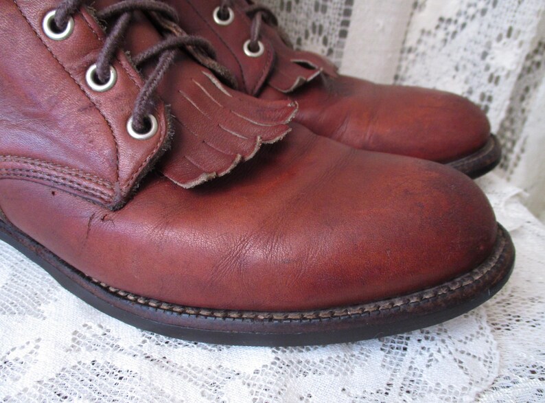 May include: Close-up of a pair of brown leather boots. The boots have a lace-up design with metal eyelets and decorative fringe on the top. The soles are dark brown with visible stitching. The leather has a worn, textured appearance.