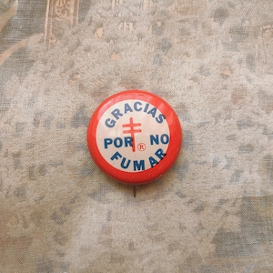 Könnte beinhalten: Roter und blauer Knopf mit dem Text "GRACIAS POR NO FUMAR" und einem Kreuzsymbol. Der Knopf hat ein eingetragenes Warenzeichen.