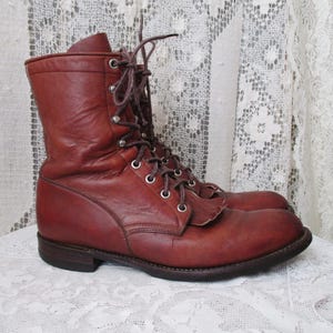 May include: A pair of brown leather work boots with laces and metal eyelets. The boots have a mid-calf height and a sturdy sole. The leather has a worn appearance, suggesting vintage or used condition. The boots are displayed on a white lace surface.