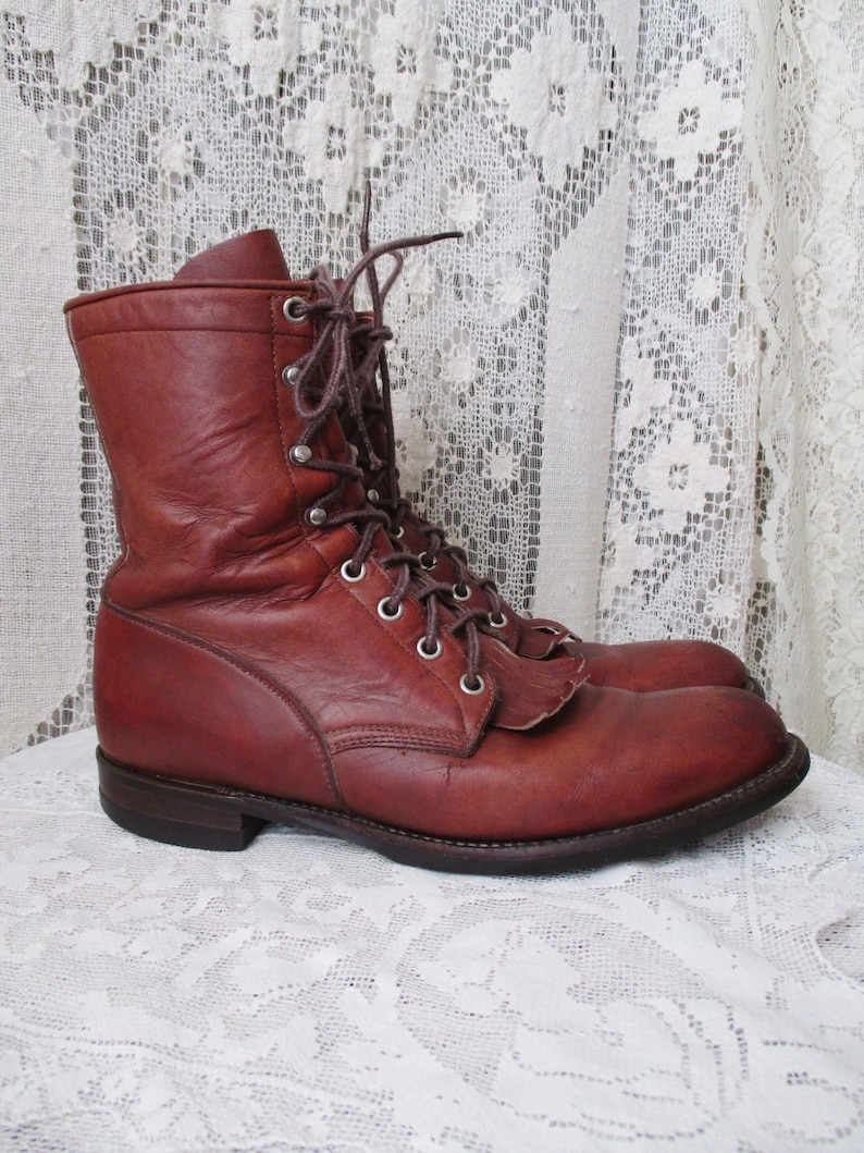 May include: A pair of brown leather work boots with laces and metal eyelets. The boots have a mid-calf height and a sturdy sole. The leather has a worn appearance, suggesting vintage or used condition. The boots are displayed on a white lace surface.