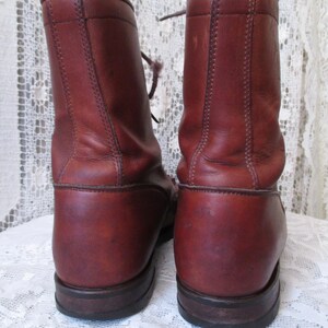 May include: A pair of reddish-brown leather boots with laces. The boots have a rounded toe and a sturdy sole. The leather shows signs of wear, adding to their vintage appeal. The boots are set against a white lace background.