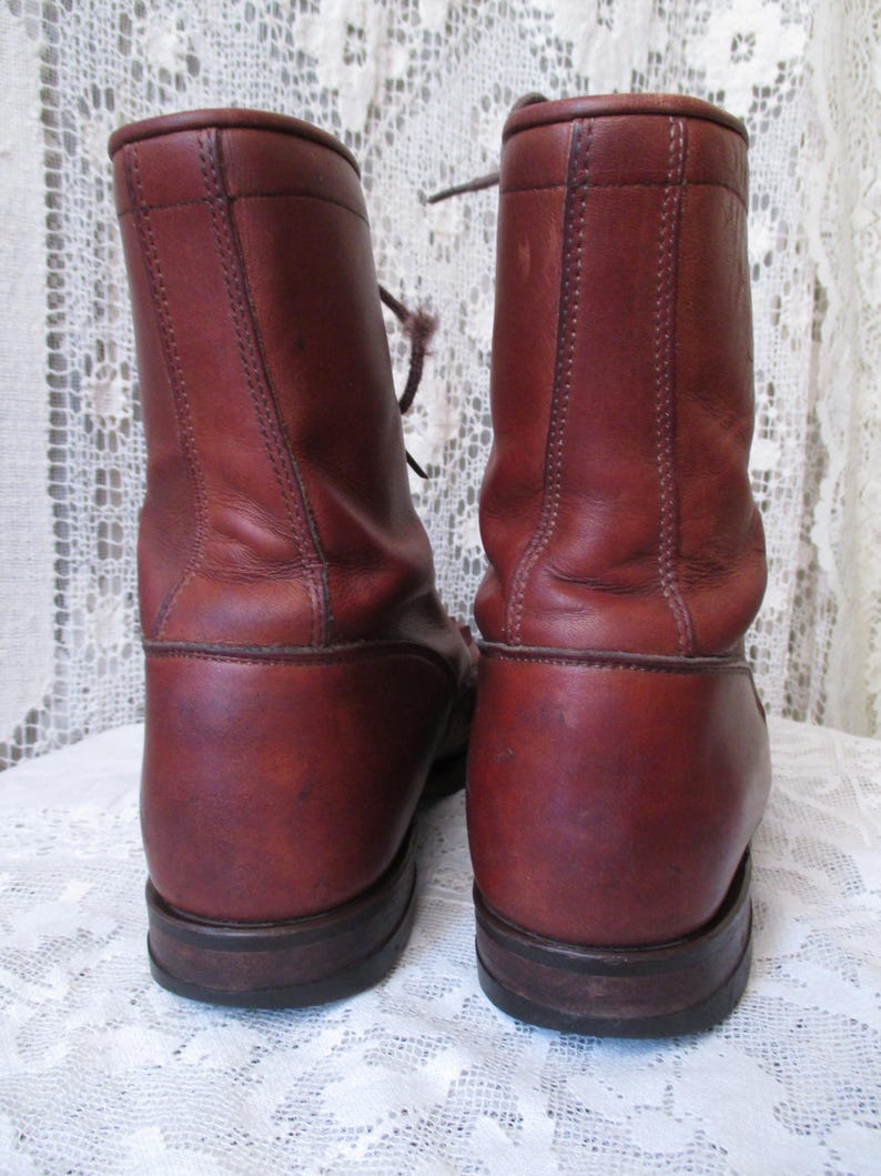 May include: A pair of reddish-brown leather boots with laces. The boots have a rounded toe and a sturdy sole. The leather shows signs of wear, adding to their vintage appeal. The boots are set against a white lace background.