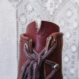 May include: Close-up of a burgundy leather boot with a lace-up design. The boot features metal eyelets and a brown lace. The top of the boot shows wear and tear, adding to its vintage aesthetic.
