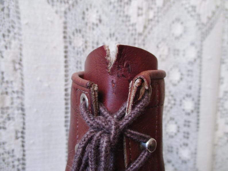 May include: Close-up of a burgundy leather boot with a lace-up design. The boot features metal eyelets and a brown lace. The top of the boot shows wear and tear, adding to its vintage aesthetic.
