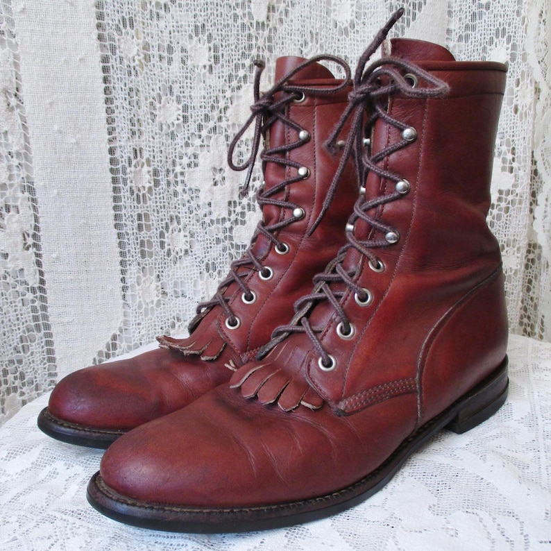 May include: A pair of burgundy leather boots with a lace-up design. The boots have a mid-calf height, a round toe, and a small fringe detail on the front. The boots are displayed on a white lace surface.