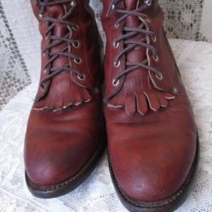 May include: A pair of reddish-brown leather boots with laces and fringe detailing. The boots have a worn appearance, with visible creases and scuffs. The laces are brown, and the soles are dark brown.
