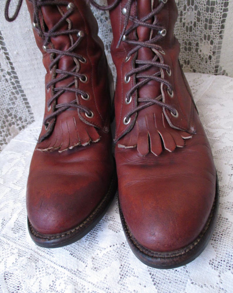 May include: A pair of reddish-brown leather boots with laces and fringe detailing. The boots have a worn appearance, with visible creases and scuffs. The laces are brown, and the soles are dark brown.