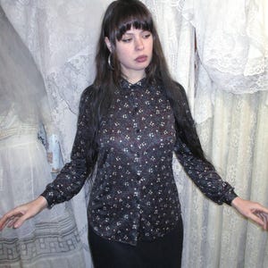 May include: A long-sleeved button-down shirt with a dark background and a small, repeating floral pattern in white and burgundy. The shirt is paired with a black skirt. The background features white lace curtains.