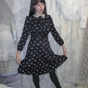 Pode incluir: Um vestido preto com um padrão de folhas brancas e uma gola de renda branca. O vestido de manga comprida tem uma frente abotoada e uma saia rodada. A pessoa na imagem está usando meias pretas e sapatilhas pretas.