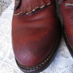 May include: Close-up of a pair of reddish-brown leather boots. The boots have a fringe detail on the top and a stitched sole. The leather shows signs of wear and tear, adding to their vintage appeal.
