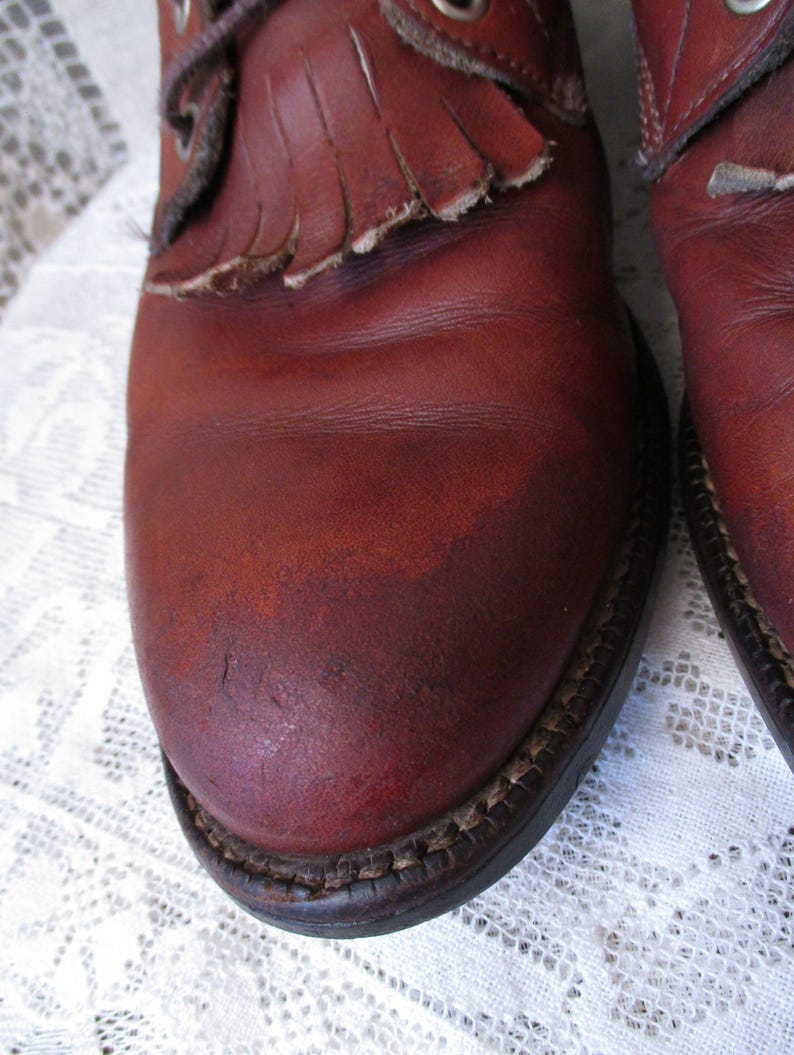 May include: Close-up of a pair of reddish-brown leather boots. The boots have a fringe detail on the top and a stitched sole. The leather shows signs of wear and tear, adding to their vintage appeal.