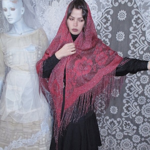 Pink Sparkly floral fringed shawl worn by a woman in all black, a long skirt and blouse, and dark hair parted to the side  with a bobby pin. A sixties mannequin wearing layers of antique white clothing is in the background, along with white lace.