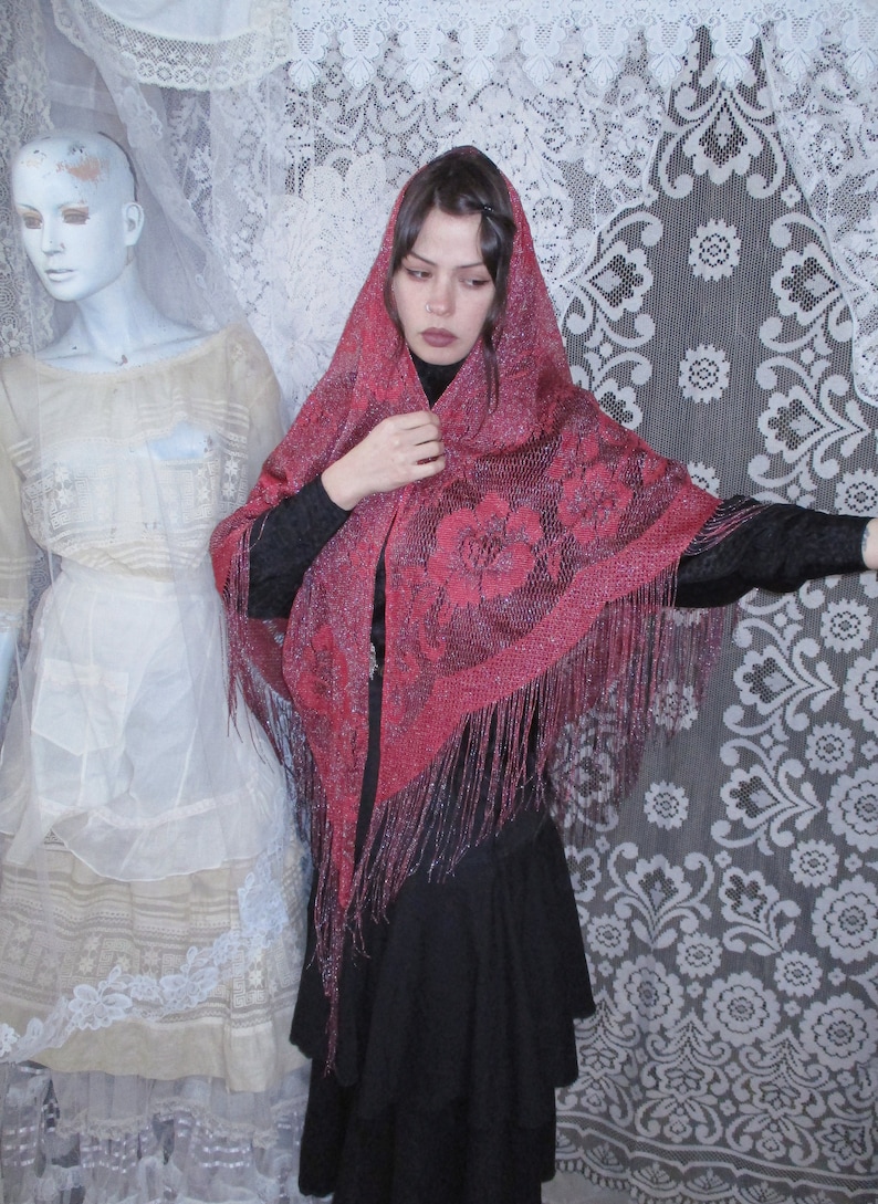 Pink Sparkly floral fringed shawl worn by a woman in all black, a long skirt and blouse, and dark hair parted to the side  with a bobby pin. A sixties mannequin wearing layers of antique white clothing is in the background, along with white lace.