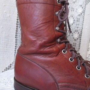 May include: A close-up of a brown leather boot with a lace-up design. The boot features a high ankle, metal eyelets, and a sturdy sole. The leather has a worn appearance, suggesting durability and age. The laces are brown.