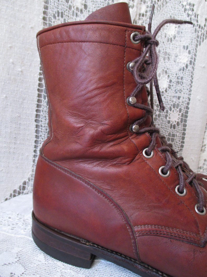 May include: A close-up of a brown leather boot with a lace-up design. The boot features a high ankle, metal eyelets, and a sturdy sole. The leather has a worn appearance, suggesting durability and age. The laces are brown.