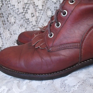 May include: Close-up of a pair of burgundy leather boots. The boots have a lace-up design with metal eyelets and decorative fringe. The soles are dark brown with visible stitching. The boots are on a white lace surface.