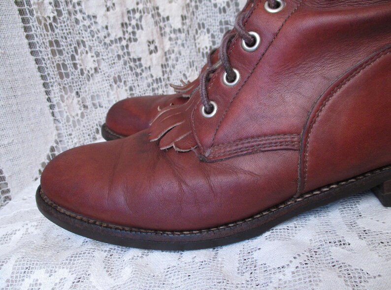 May include: Close-up of a pair of burgundy leather boots. The boots have a lace-up design with metal eyelets and decorative fringe. The soles are dark brown with visible stitching. The boots are on a white lace surface.