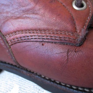 May include: Close-up of a brown leather boot. The boot shows detailed stitching, a metal eyelet, and signs of wear. The leather has a rich, reddish-brown hue, with visible texture and slight imperfections.