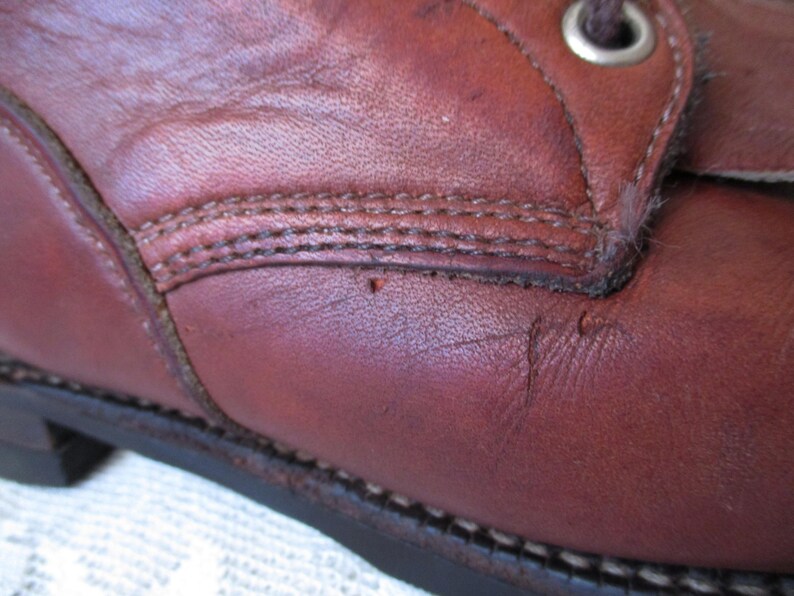 May include: Close-up of a brown leather boot. The boot shows detailed stitching, a metal eyelet, and signs of wear. The leather has a rich, reddish-brown hue, with visible texture and slight imperfections.