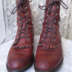 May include: A pair of reddish-brown leather boots with laces and metal eyelets. The boots have a fringe detail at the top of the foot and a dark brown sole. The laces are tied in a bow at the top.