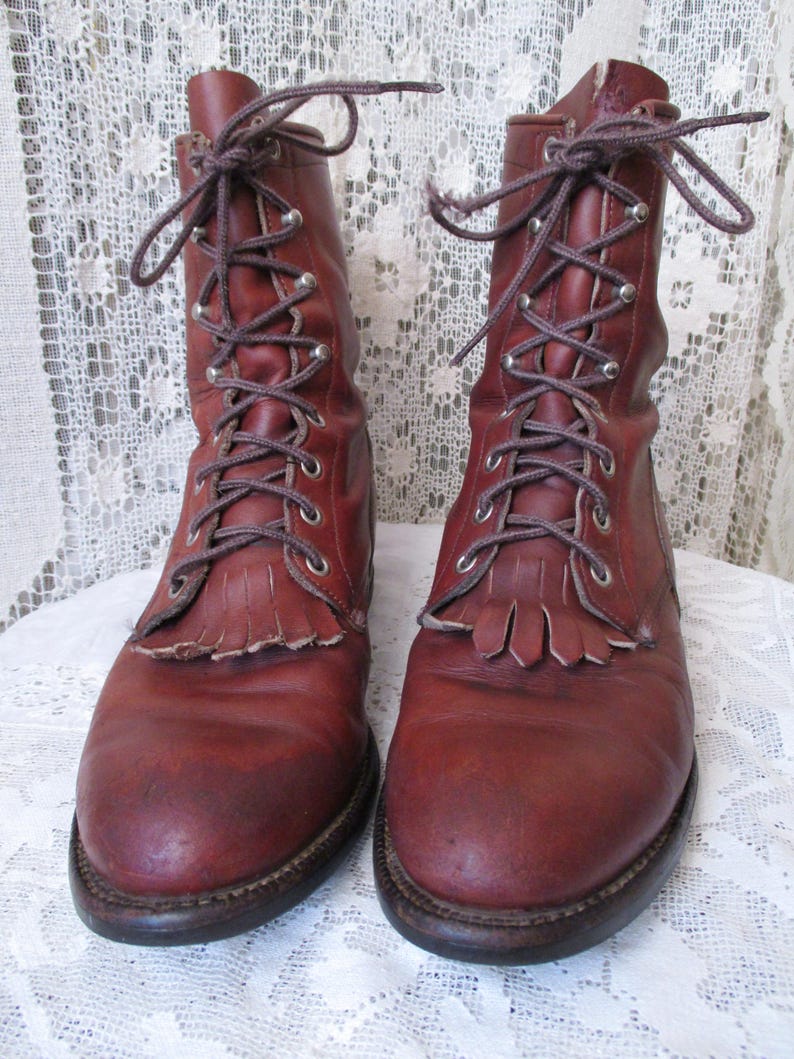 May include: A pair of reddish-brown leather boots with laces and metal eyelets. The boots have a fringe detail at the top of the foot and a dark brown sole. The laces are tied in a bow at the top.