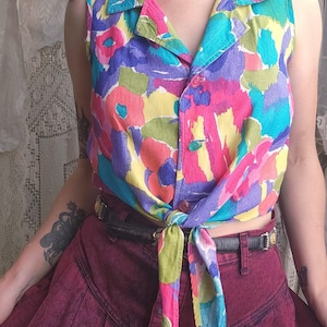 May include: A sleeveless button-down shirt featuring a vibrant, multi-colored abstract floral print in shades of pink, yellow, blue, and green. The shirt has a collared neckline and is tied at the waist, paired with a dark maroon pleated skirt.