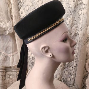 Puede incluir: Un sombrero negro con una corona rectangular y un ribete dorado. El sombrero tiene una cinta azul marino oscuro que cuelga por la espalda. El sombrero se exhibe en una cabeza de maniquí.