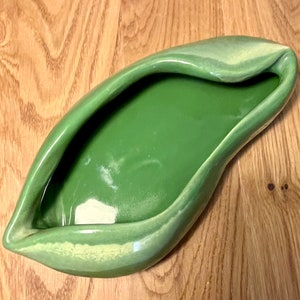 May include: A green ceramic bowl shaped like a leaf. The bowl has a smooth, glossy finish and a slightly rippled edge.
