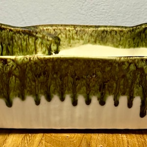 May include: A green and white ceramic planter with a unique, wavy design. The planter has a wide, rectangular base and a slightly curved top.
