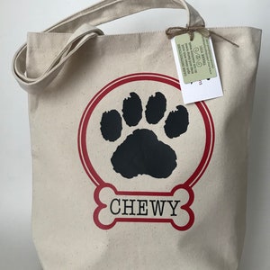 May include: A beige canvas tote bag with red and black paw print design. The bag has a red bone with the name "CHEWY" printed on it. The bag has a tag attached with care instructions.