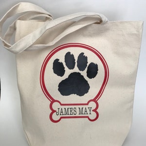 May include: A white canvas tote bag with red and black paw print design. The bag has red and black bone-shaped design with the text "JAMES MAY" inside.