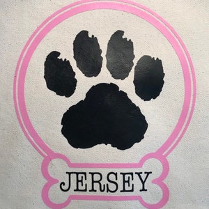 May include: A white canvas tote bag with a pink paw print design. The paw print is surrounded by a pink circle. The text "JERSEY" is written in black letters on a pink bone shape.