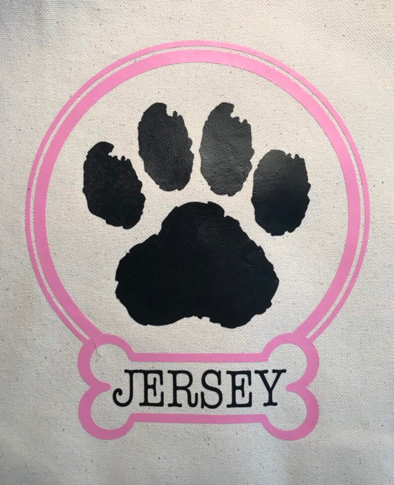 May include: A white canvas tote bag with a pink paw print design. The paw print is surrounded by a pink circle. The text "JERSEY" is written in black letters on a pink bone shape.