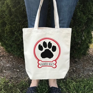 May include: A white canvas tote bag with a red circle and black paw print design. The text "JAMES MAY" is printed below the paw print.