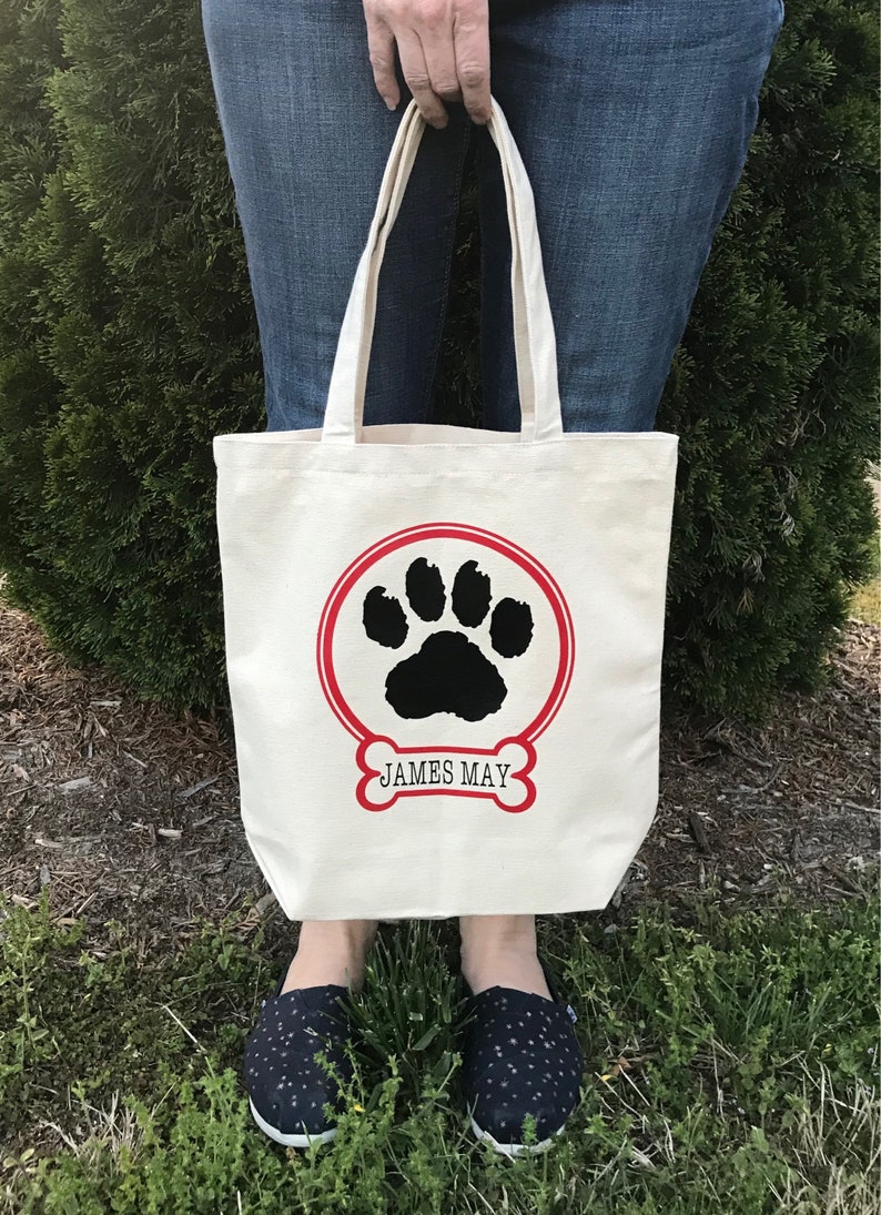 May include: A white canvas tote bag with a red circle and black paw print design. The text "JAMES MAY" is printed below the paw print.