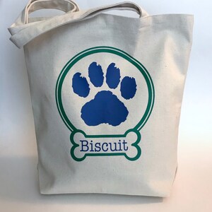 May include: A white canvas tote bag with a green circle and a blue paw print. The text "Biscuit" is written in a green bone shape.