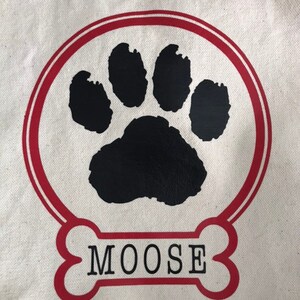 May include: A white canvas tote bag with a red circle and a black paw print inside. The circle has a red outline. Below the circle is a red bone with the word "MOOSE" in black text.