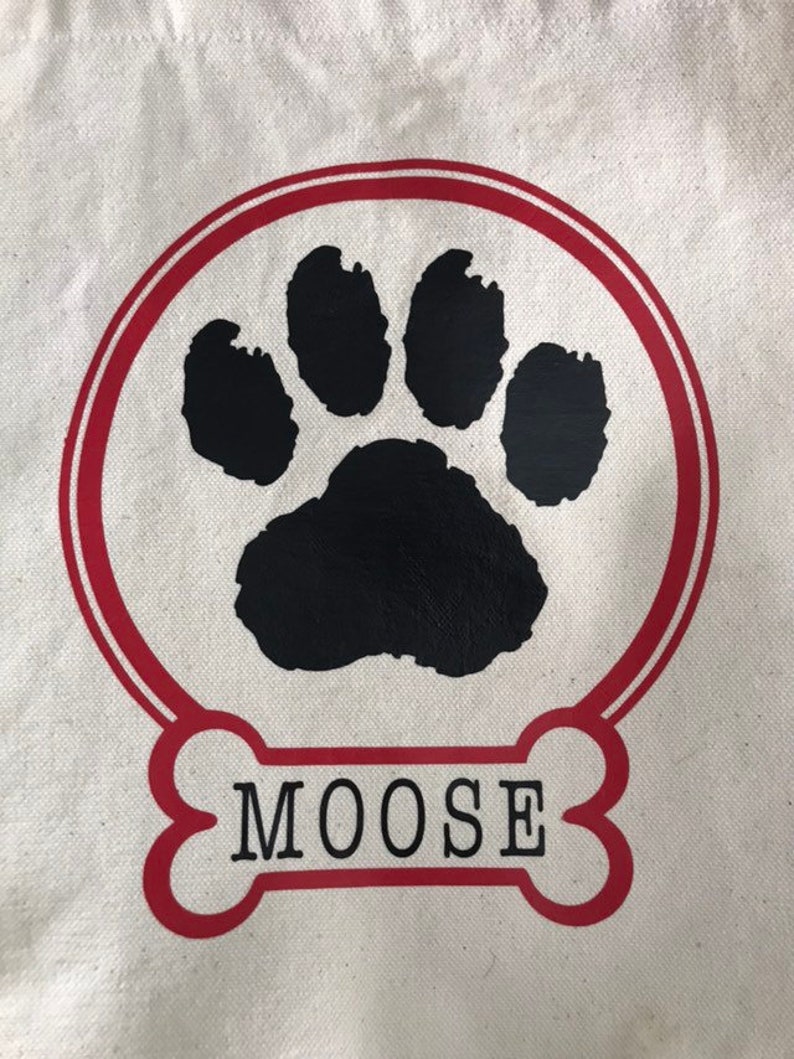 May include: A white canvas tote bag with a red circle and a black paw print inside. The circle has a red outline. Below the circle is a red bone with the word "MOOSE" in black text.