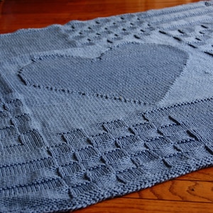 May include: A blue knitted blanket with a large heart design in the center. The blanket is made with a simple stitch pattern and has a soft, textured feel.