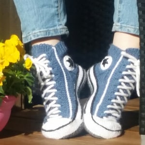 May include: A pair of blue knitted high-top sneaker socks with white laces, soles, and star patches. The socks are designed to resemble classic sneakers. The image shows the socks being worn, with the wearer's legs visible above the ankles.