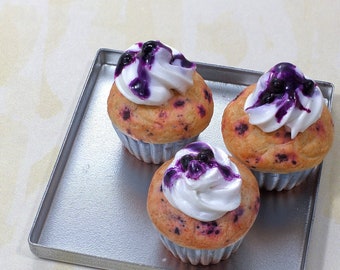 May include: Three mini cupcakes with white frosting and blueberry topping on a silver tray.