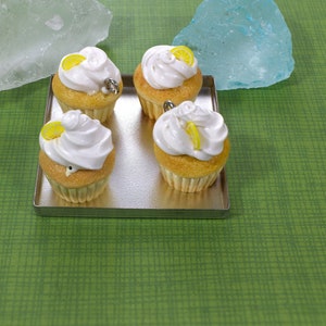 May include: Four miniature lemon cupcakes with white frosting and a lemon slice on top, displayed on a silver tray. The cupcakes are on a green background.