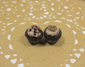 May include: Two miniature chocolate cupcakes with brown and white frosting. One cupcake is topped with chocolate sprinkles and the other with brown sprinkles.