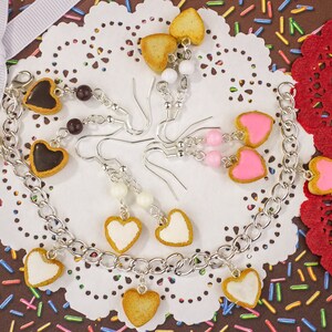 May include: A collection of handmade jewelry featuring heart-shaped charms. The jewelry includes a silver chain bracelet and several pairs of earrings. The heart charms have various colors, including brown, white, and pink, with a biscuit-like border. The background is a brown surface with colorful sprinkles.