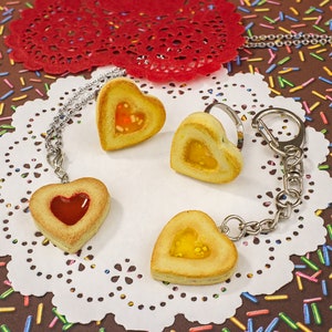 May include: Heart-shaped pendants and keychains with a cookie-like appearance. The pendants and keychains have a filling of red and yellow jelly. The items are on a white doily, with a red paper doily and a brown background with colorful sprinkles.