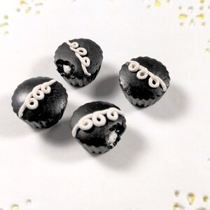 May include: Four miniature black cupcakes with white frosting swirls. The cupcakes are on a white background with a lace pattern.