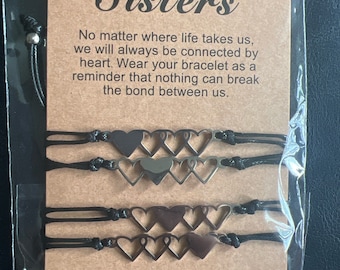 Può includere: Set di quattro braccialetti in corda nera con ciondoli a forma di cuore argentati. I ciondoli sono cuori aperti con un cuore più piccolo all'interno. La carta dice "Sorelle" e include un messaggio sul legame tra le sorelle.