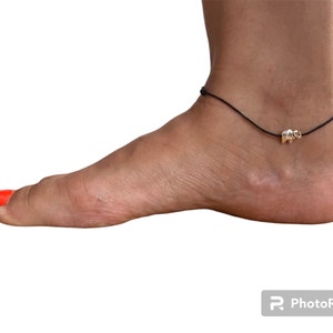 Op de afbeelding: Een zwarte enkelband met een klein zilveren olifantbedeltje. De enkelband zit om een enkel met rode nagellak.