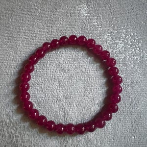 May include: A burgundy beaded bracelet. The bracelet is made of small, round beads strung together to form a circular shape. The beads have a glossy finish and are a deep, rich red colour. The bracelet is shown on a grey surface.