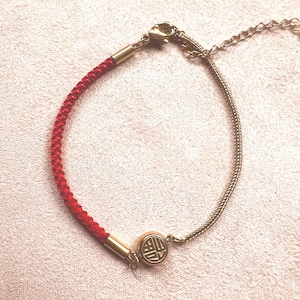 May include: A gold-toned bracelet with a red woven cord section and a delicate chain. The bracelet features a circular charm with an embossed design. The clasp and connecting elements are also gold-toned.